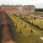 Hampton Court Palace located in London in England near arched passage covered in dry plants near green grass on lawn near stone steps and trees under bright sky in daylight