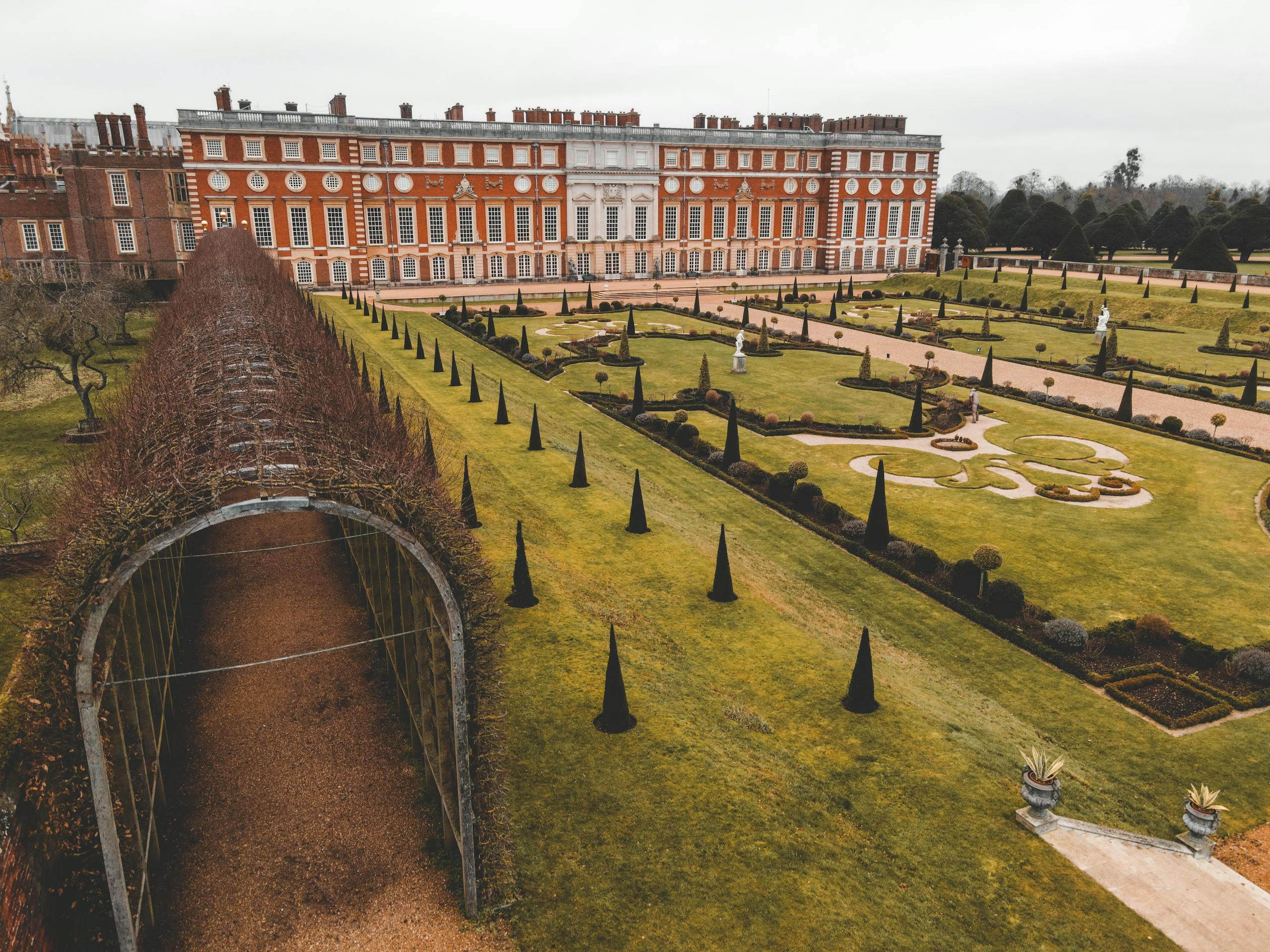Hampton Court Palace located in London in England near arched passage covered in dry plants near green grass on lawn near stone steps and trees under bright sky in daylight