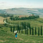 Serene view of a woman walking through picturesque Tuscan countryside with winding road and cypress trees.